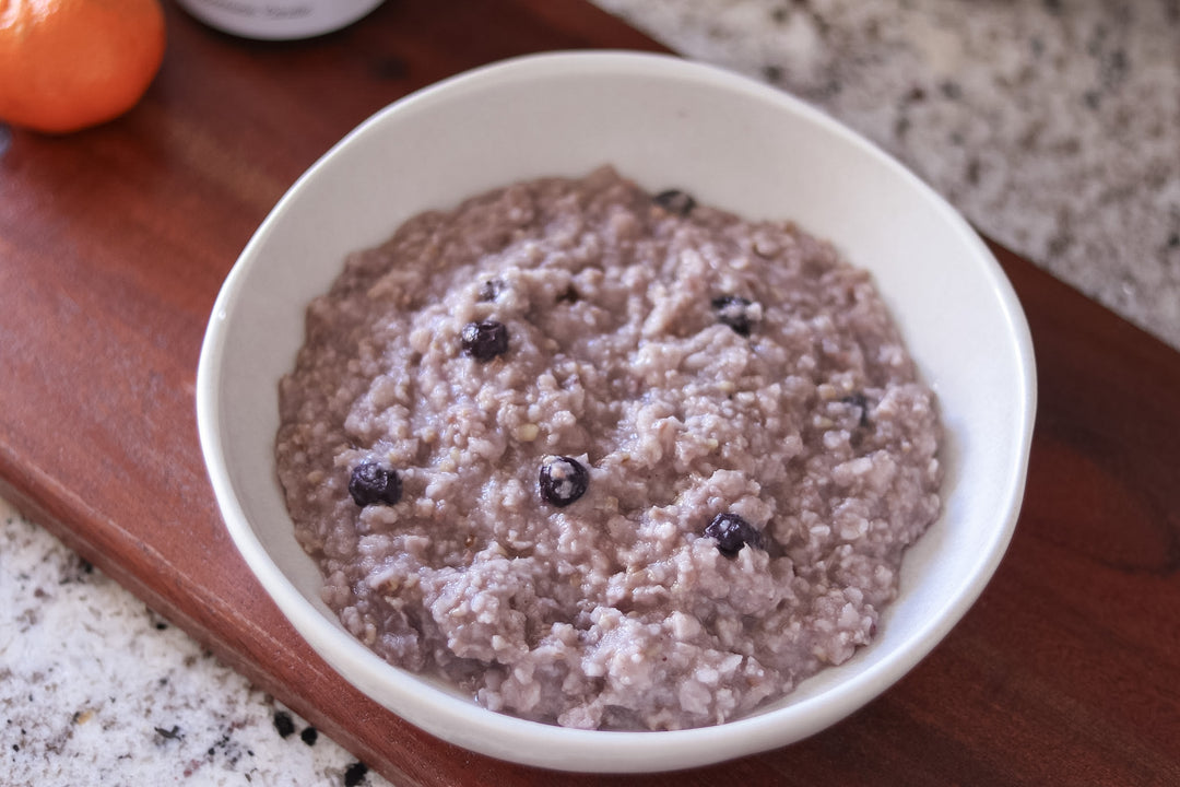 A white ceramic bowl filled with oatmeal or porridge containing dark blueberries, set on a wooden cutting board with a granite countertop in the background.