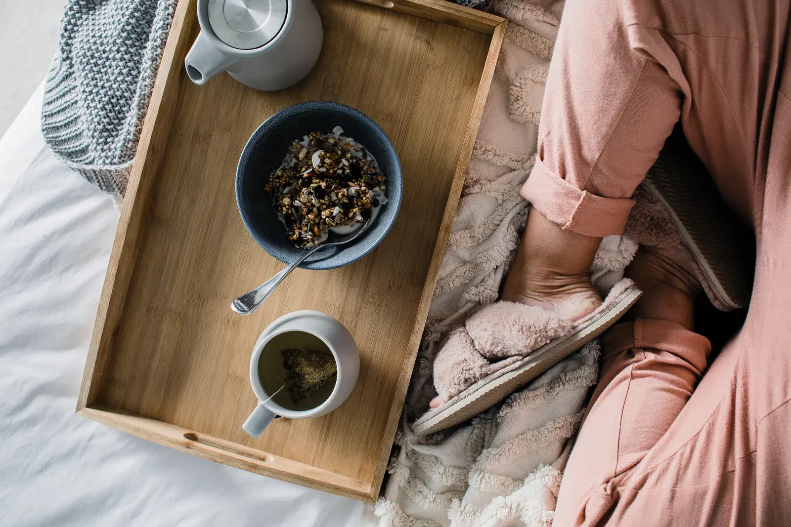 Person sitting on a bed with a wooden tray holding a cup and bowl of tea leaves.