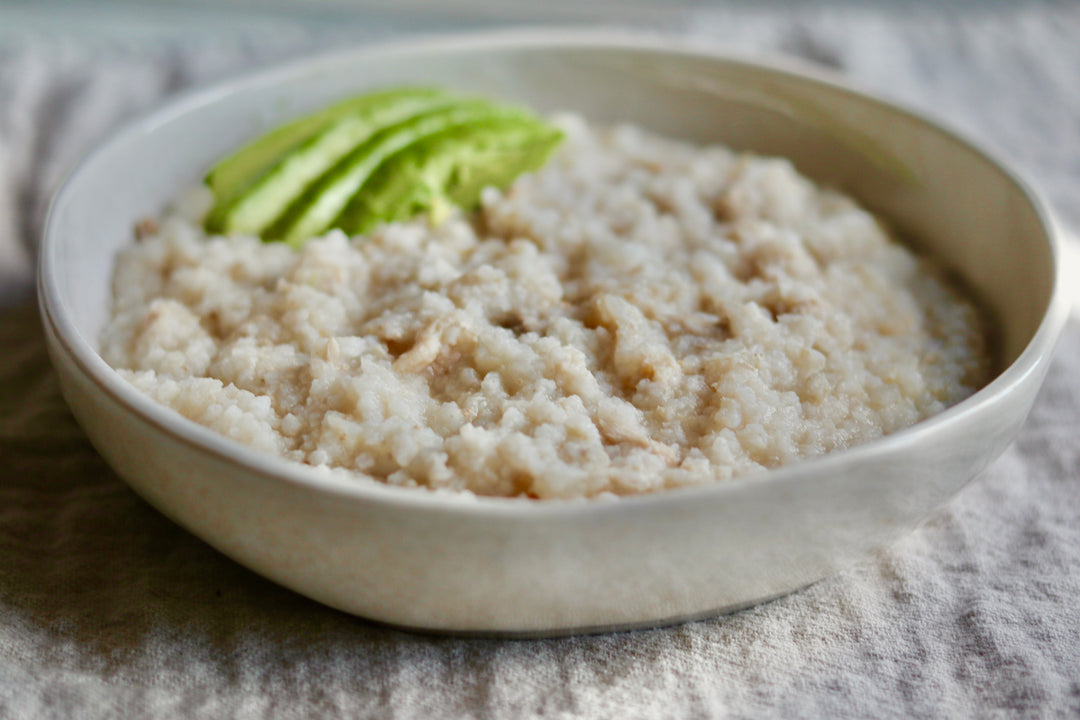 A close-up of a light beige ceramic bowl filled with cooked oatmeal or rice, garnished with a few green avocado slices, set on a textured gray cloth.