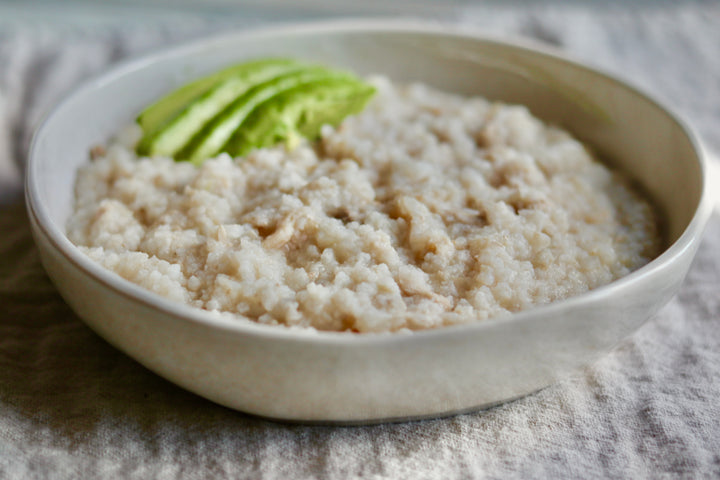 A close-up of a light beige ceramic bowl filled with cooked oatmeal or rice, garnished with a few green avocado slices, set on a textured gray cloth.