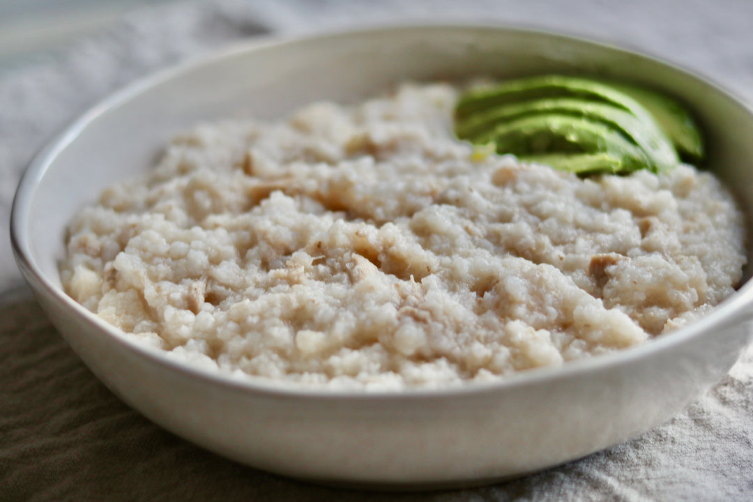 A close-up shot of a white ceramic bowl filled with oatmeal or a grain porridge, garnished with a few slices of green avocado, set on a textured gray cloth background.