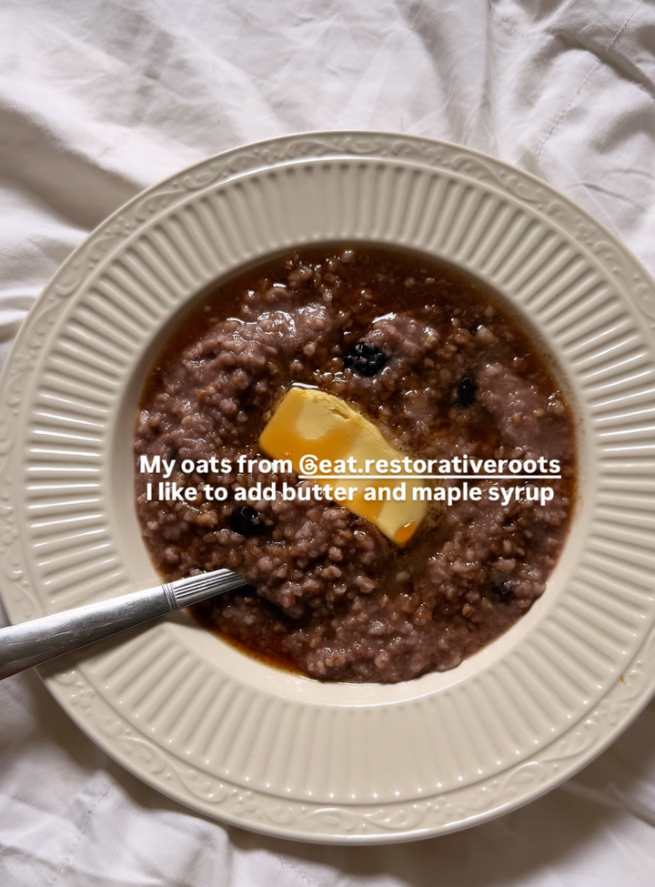 A top-down view of a bowl of oatmeal with a pat of butter on top, served on a white textured fabric background. A spoon is resting in the bowl, and there is a text overlay describing the oats.