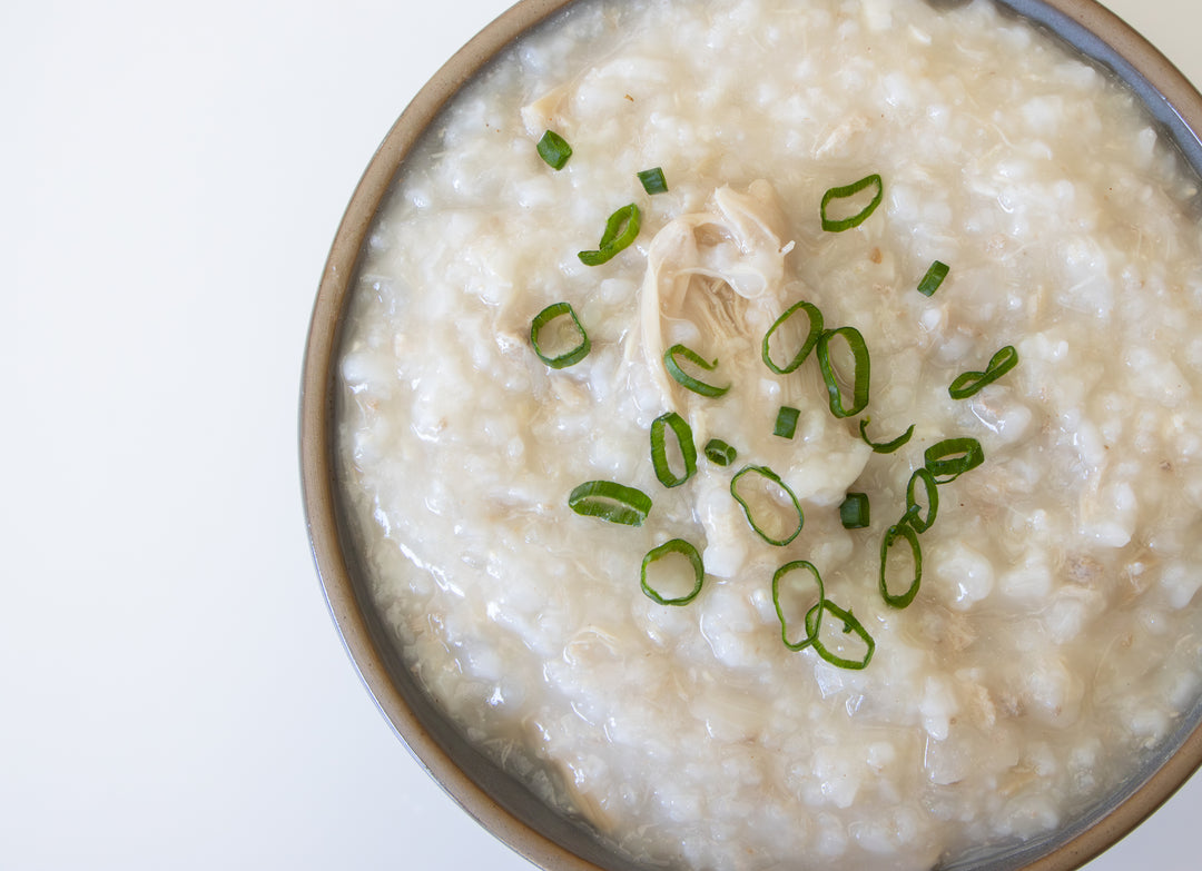 A top-down view of a bowl of creamy rice porridge or congee garnished with sliced green onions and shredded chicken, set against a clean white background.