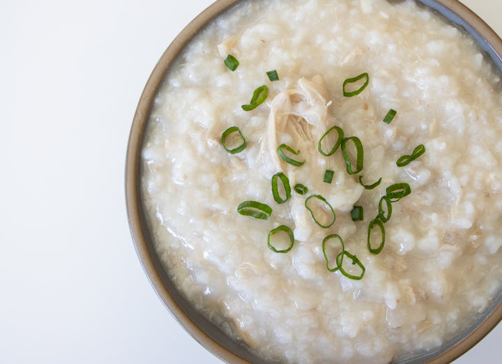 A top-down view of a bowl of creamy rice porridge or congee garnished with sliced green onions and shredded chicken, set against a clean white background.